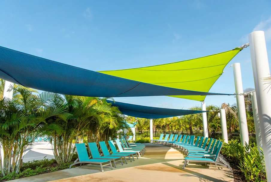 Custom shade structures with blue and green sail shades providing coverage over poolside lounge chairs, surrounded by tropical landscaping and clear blue sky.