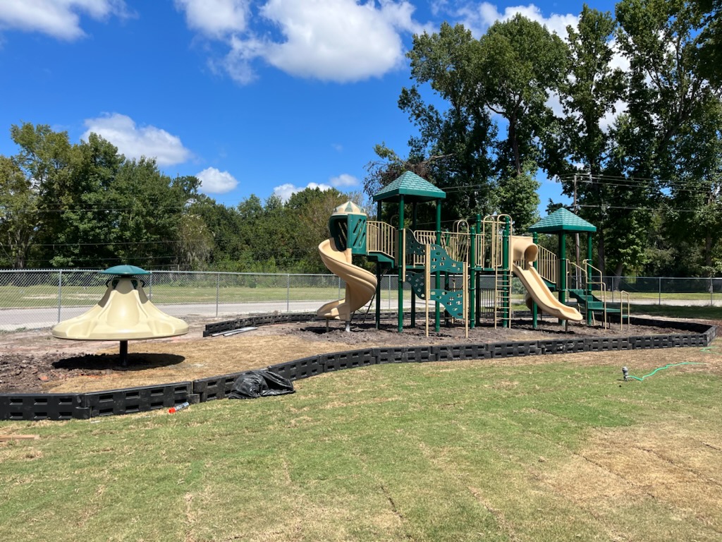 Playground equipment featuring green and tan slides, climbing structures, and a spinning feature, set in a fenced outdoor area with grass and trees, illustrating maintenance and safety for recreational spaces.