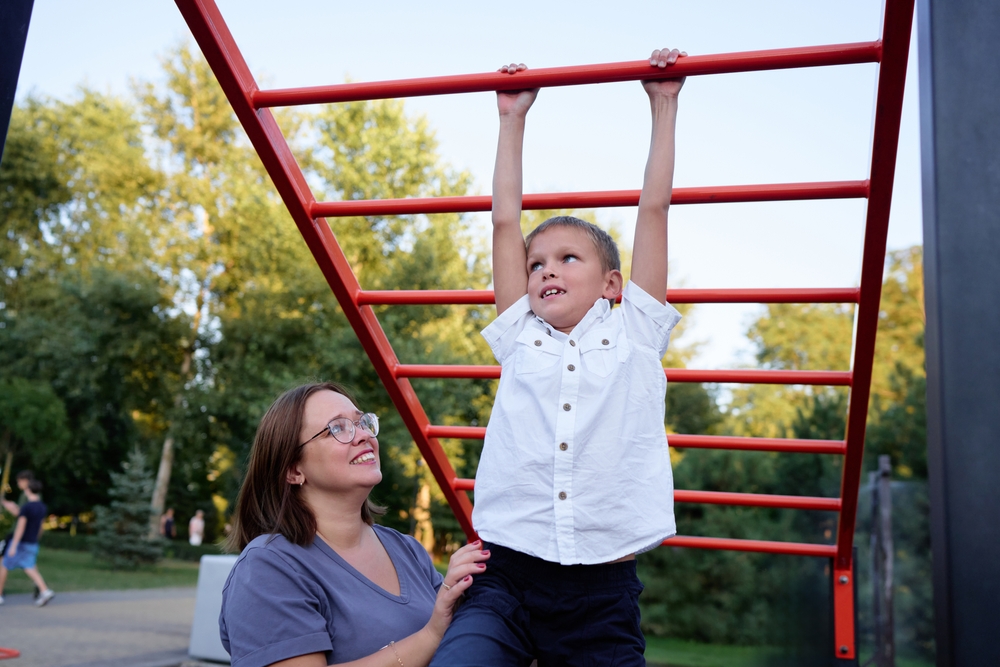 Mother supporting child on playground monkey bars, emphasizing safety and accessibility in play environments.
