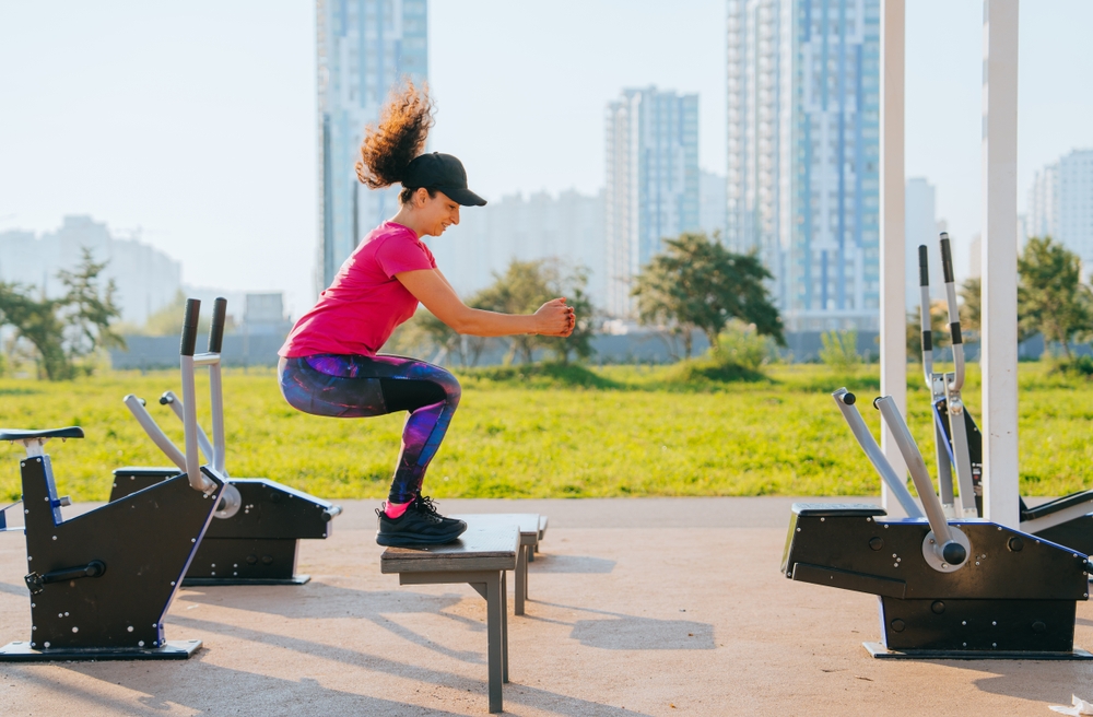Woman exercising on outdoor fitness equipment, performing a squat jump on a platform, surrounded by various gym apparatus in a community fitness park, promoting health and physical activity.